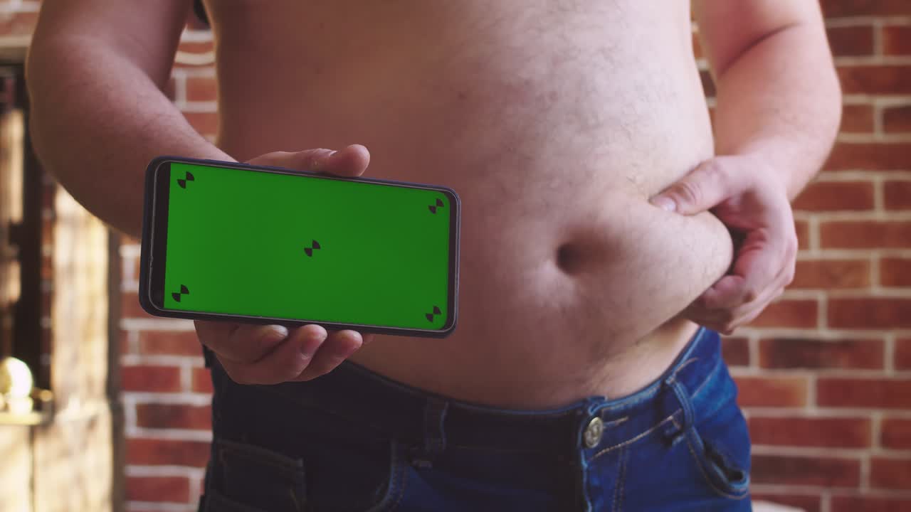 Un uomo sta in piedi, si tocca l'enorme pancia e mostra il telefono con green screen. Primo piano dell'addome. Concetto di eccesso di peso