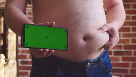 el hombre se pone de pie, toca su enorme estómago y muestra el teléfono con pantalla verde. primer plano del abdomen. concepto de exceso de peso