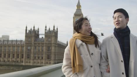 young asian couple on holiday walking across westminster bridge with houses of parliament in background