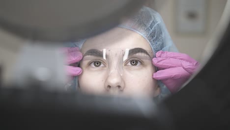 woman with brow ruler looks in mirror at tattoo in salon