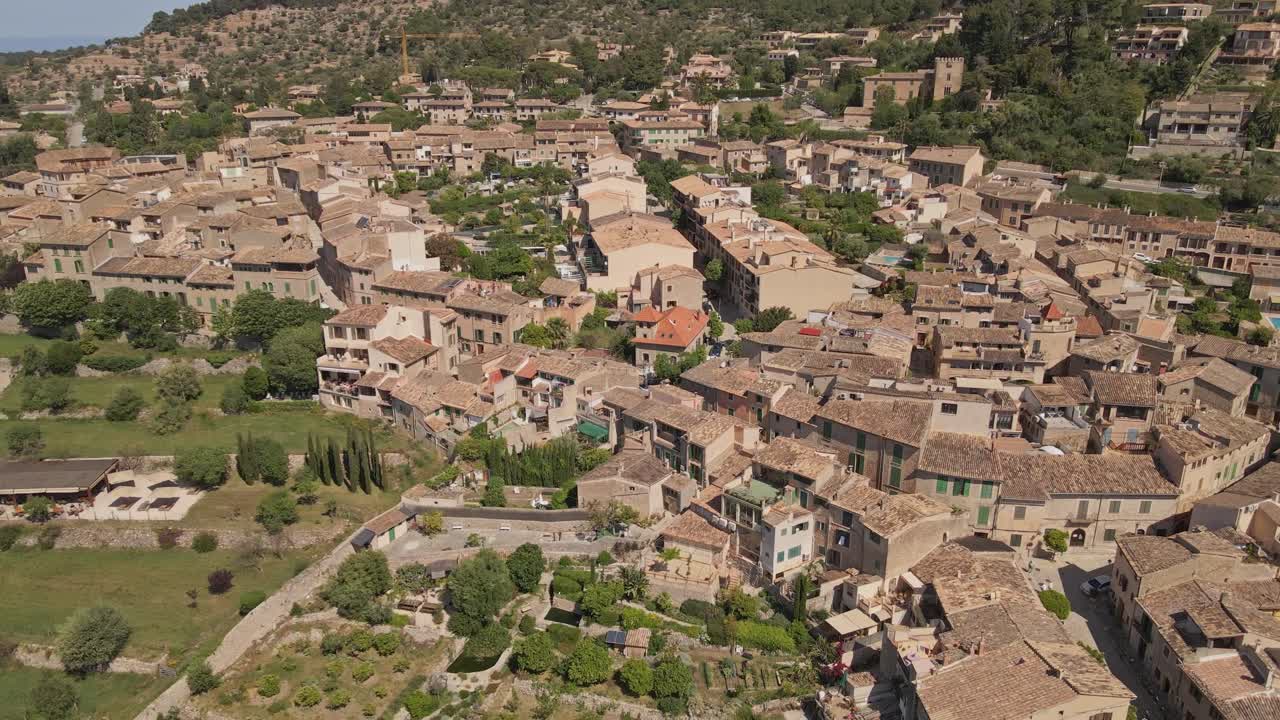 An aerial video of a traditional Mediterranean village surrounded by lush green hills and a rugged mountain backdrop. The tightly packed stone houses with terracotta roofs.