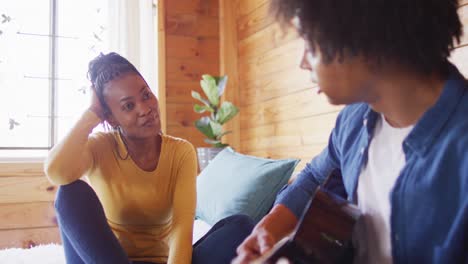 happy african american couple playing guitar, sitting on sofa in log cabin, slow motion