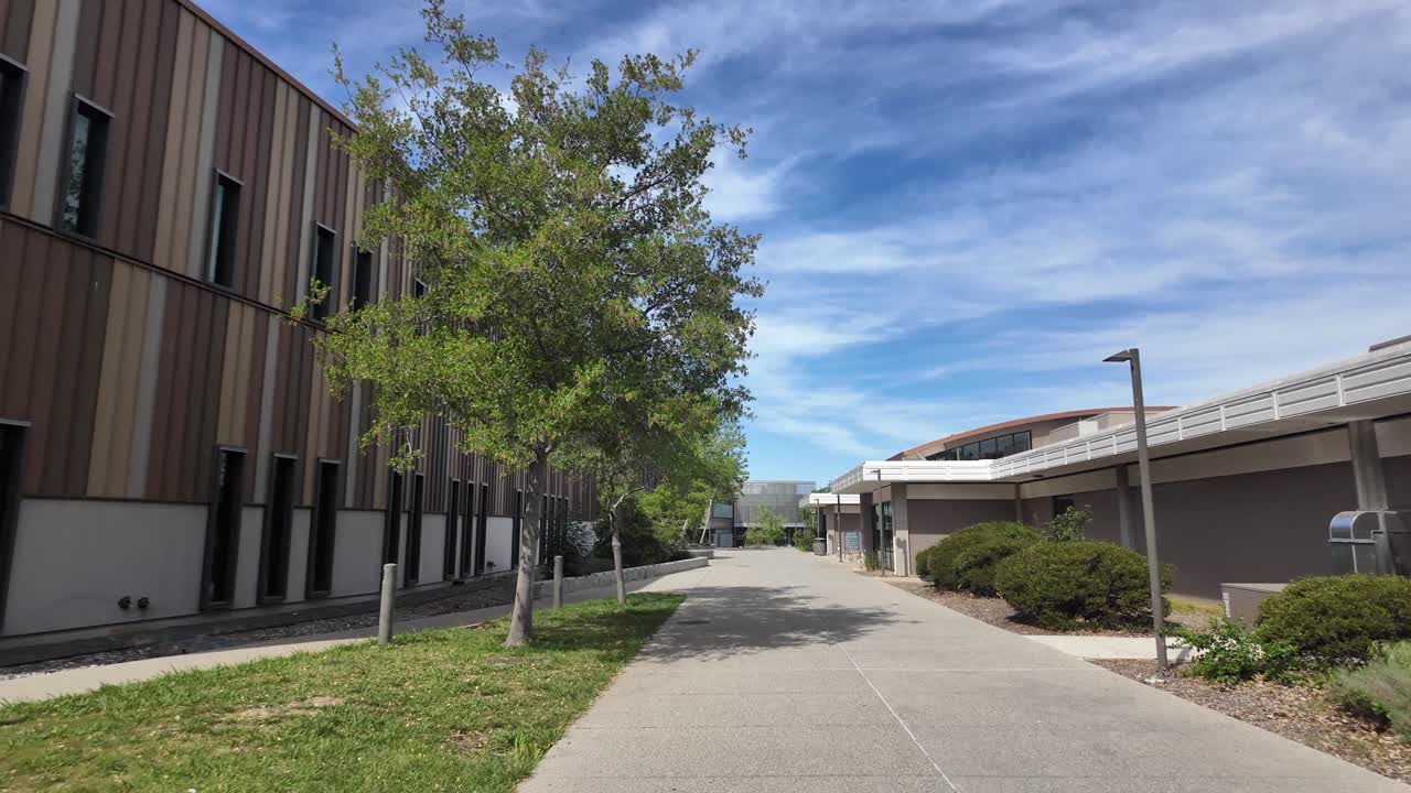 Toma en primera persona caminando lentamente por un sendero pavimentado entre modernos edificios universitarios en el campus de West Valley College en Saratoga, California, bajo un cielo azul soleado con nubes tenues.