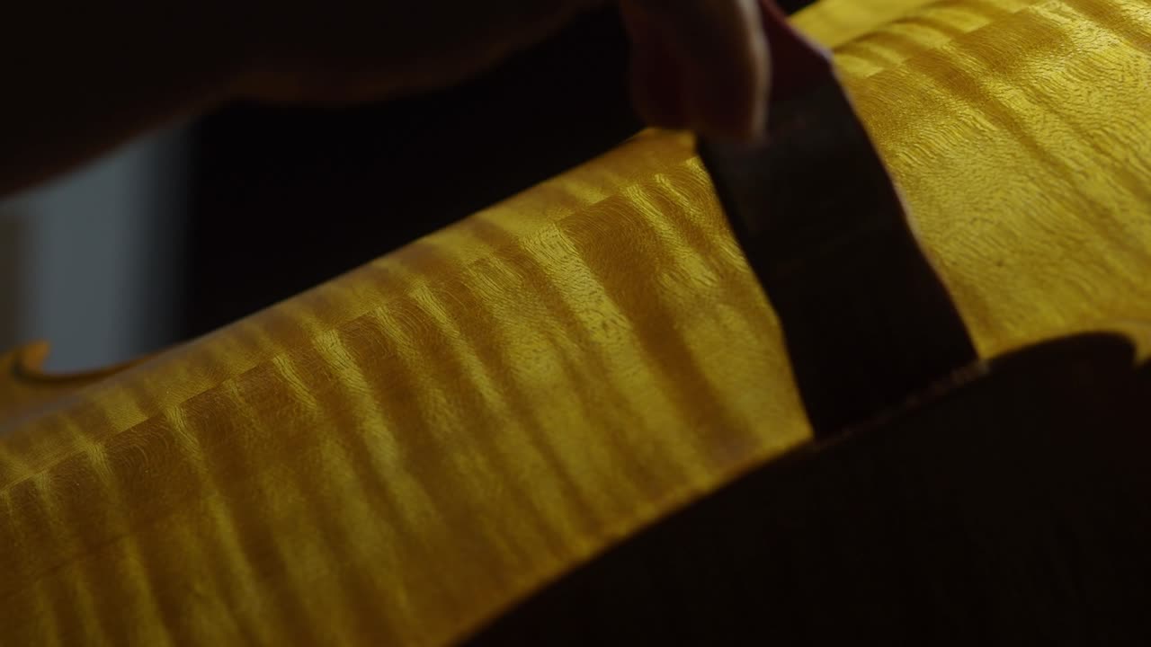 Macro view of a craftsman’s brush applying a linseed oil, mastic, and lac blend to a cello back, with vivid marbled flames and fine grain texture glowing under direct light in a traditional workshop