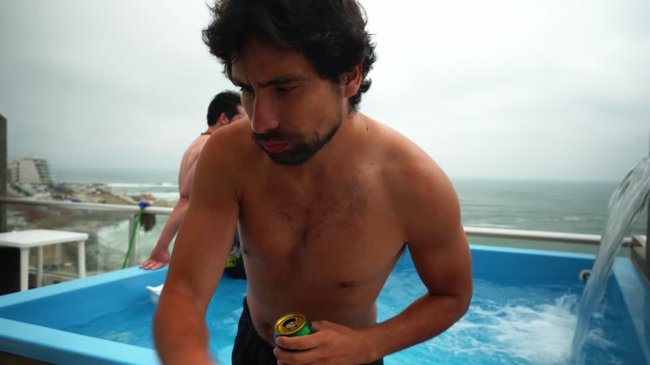 acercándose y cambiando la música con amigos sentados en el borde de una piscina en una casa con vistas a la playa con el agua cayendo a su lado y charlando y bebiendo cerveza enlatada