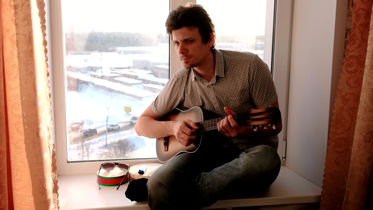 Playing a musical instrument. Man is playing the guitar sitting in windowsill.