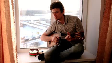 playing a musical instrument. man is playing the guitar sitting in windowsill.