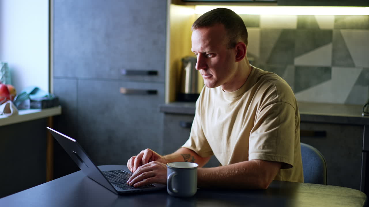 Busy Caucasian man working on his laptop in the kitchen. Focused freelancer looks at the screen and types on keyboard.