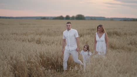 Junges-Elternpaar-Mit-Mädchen,-Die-Sich-An-Den-Händen-Halten-Und-Bei-Sonnenuntergang-Durch-Das-Weizenfeld-Laufen.-Glückliche-Familie-Joggt-Auf-Der-Gerstenwiese-Und-Genießt-Gemeinsam-Die-Natur.-Zeitlupe