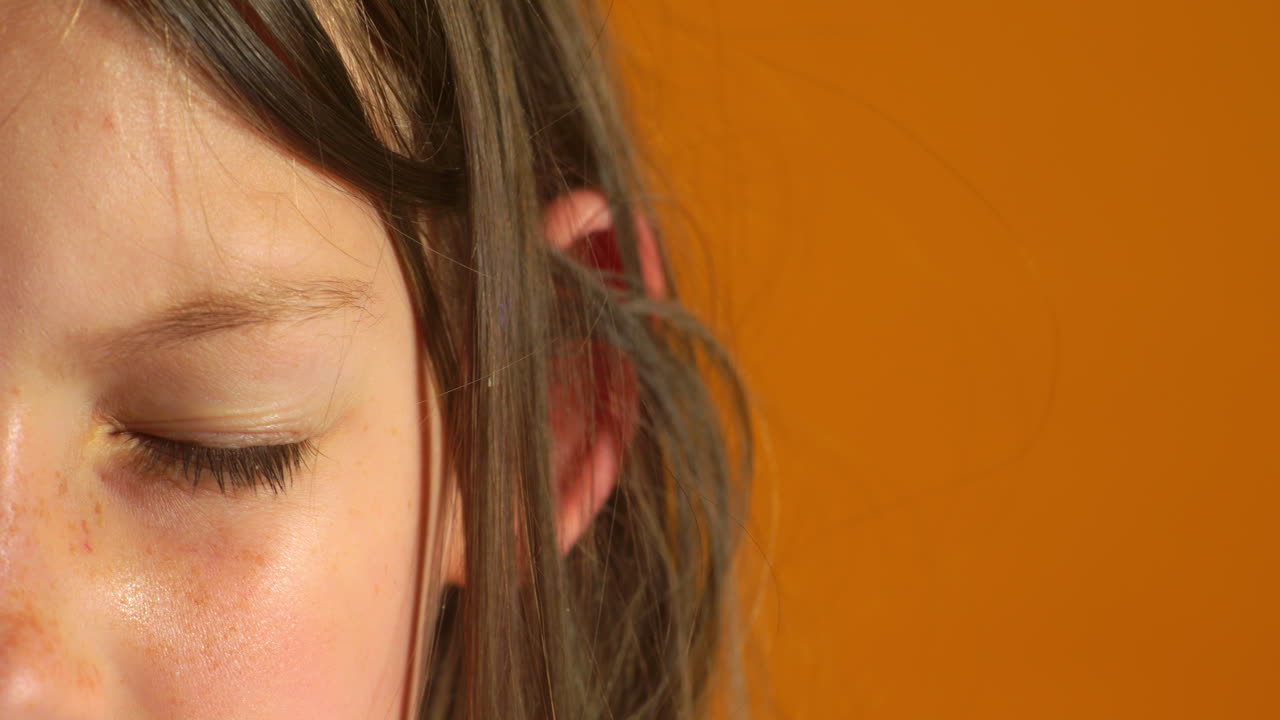 A soft close-up of a child’s face shows one eye slowly opening in a calm, relaxed moment against a warm orange background, capturing a simple and natural expression of curiosity or attention.