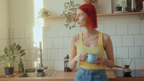 young woman standing in kitchen, holding coffee cup and looking through window