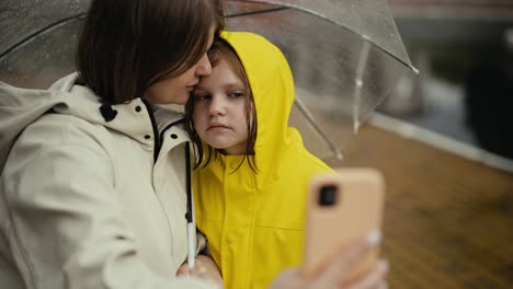 Happy-blonde-woman-in-a-white-jacket-takes-a-selfie-with-her-teenage-daughter-in-a-yellow-jacket-and-holds-an-umbrella-while-walking-after-the-rain-in-the-park