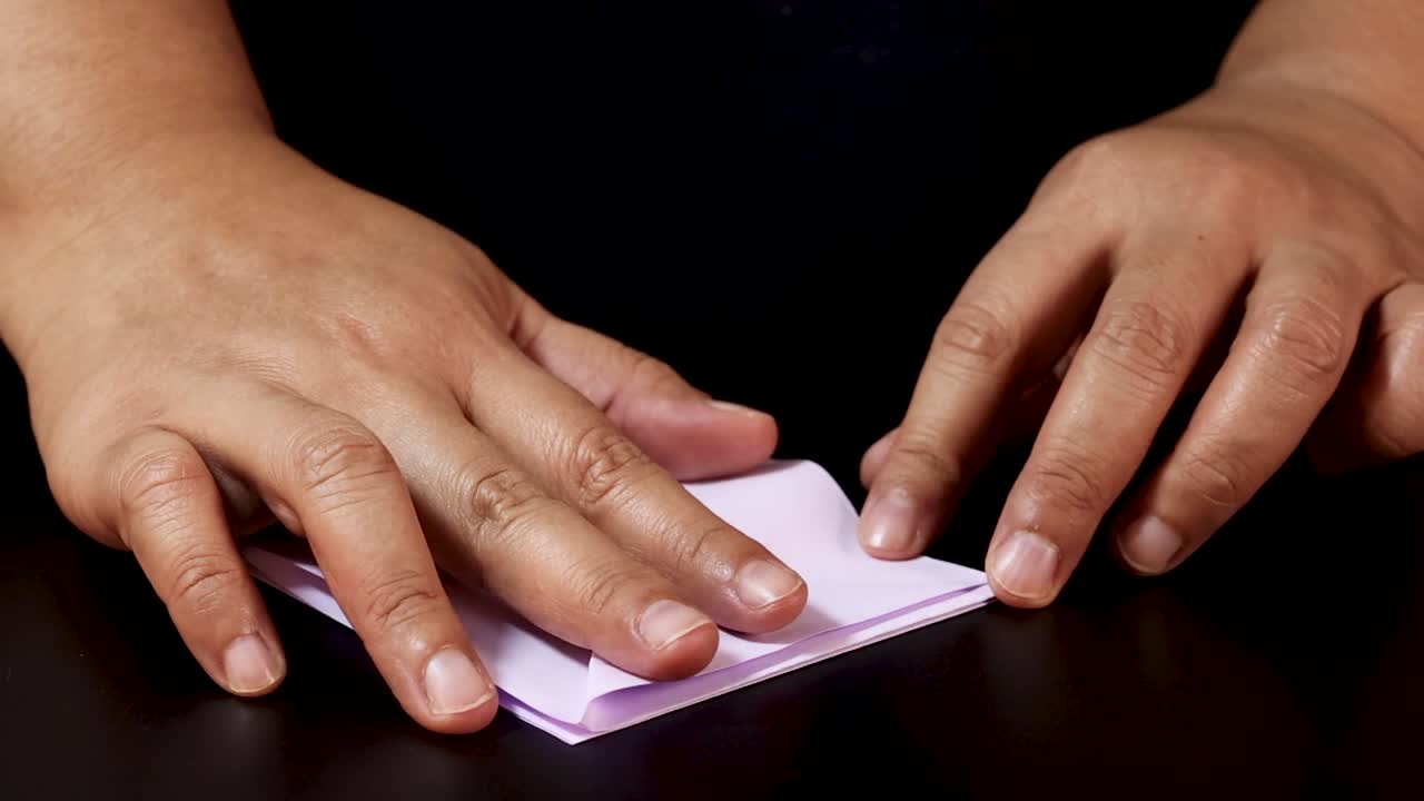 Adult hands carefully fold a square of purple paper into origami shapes against black background