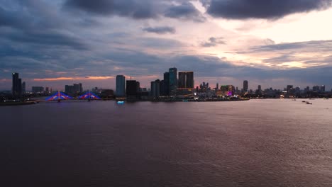 aerial footage of phnom penh at dusk, showcasing the serene mekong river and city skyline under a vibrant, colorful sky. the urban landscape glows invitingly in the soft evening light