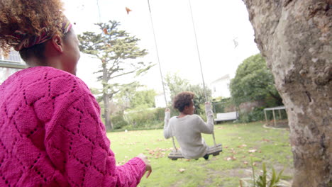 happy african american mother pushing her son on tree swing in garden, slow motion