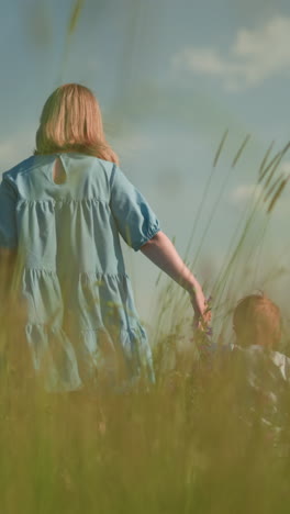 back view of a woman in a flowing blue gown walking with her two young sons, both in white shirts, through a lush green grass field under a clear blue sky with scattered clouds