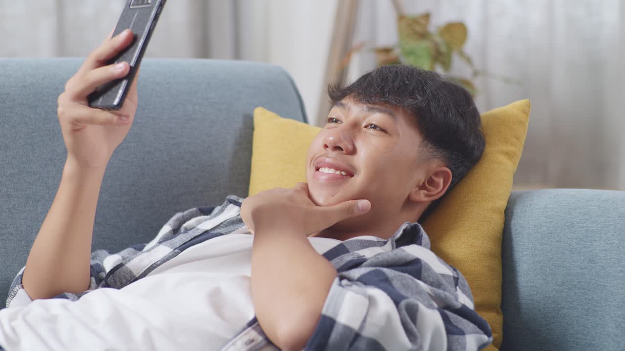스마트폰을 사용하는 아시아 십대 소년이 집 안의 거실에서 소파에 누워서 셀피를 찍고 있다. 웃고, 평화를 보여주고, 엄지손가락을 높이 들고, 심장을 움직인다.