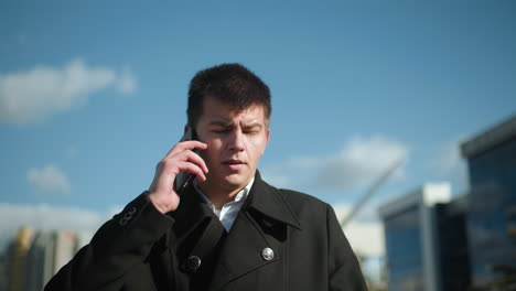 businessman wearing black coat with metallic buttons, on phone call outdoors under bright sky, looking contemplatively with blurred modern buildings and greenery in background