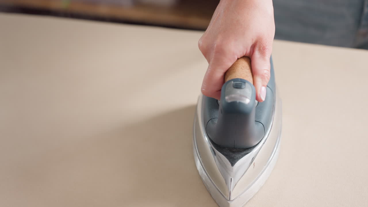 Hand view of hand holding pressing iron moving round cloth spread on ironing table as steam comes out, focusing on skillful technique, delivering neat finishing, refining garment details