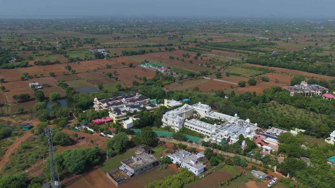 Ripresa cinematografica con drone di un complesso alberghiero di lusso bianco circondato da alberi verdi e campi agricoli nell'India rurale. Vista dall'alto che mostra architettura e paesaggio.