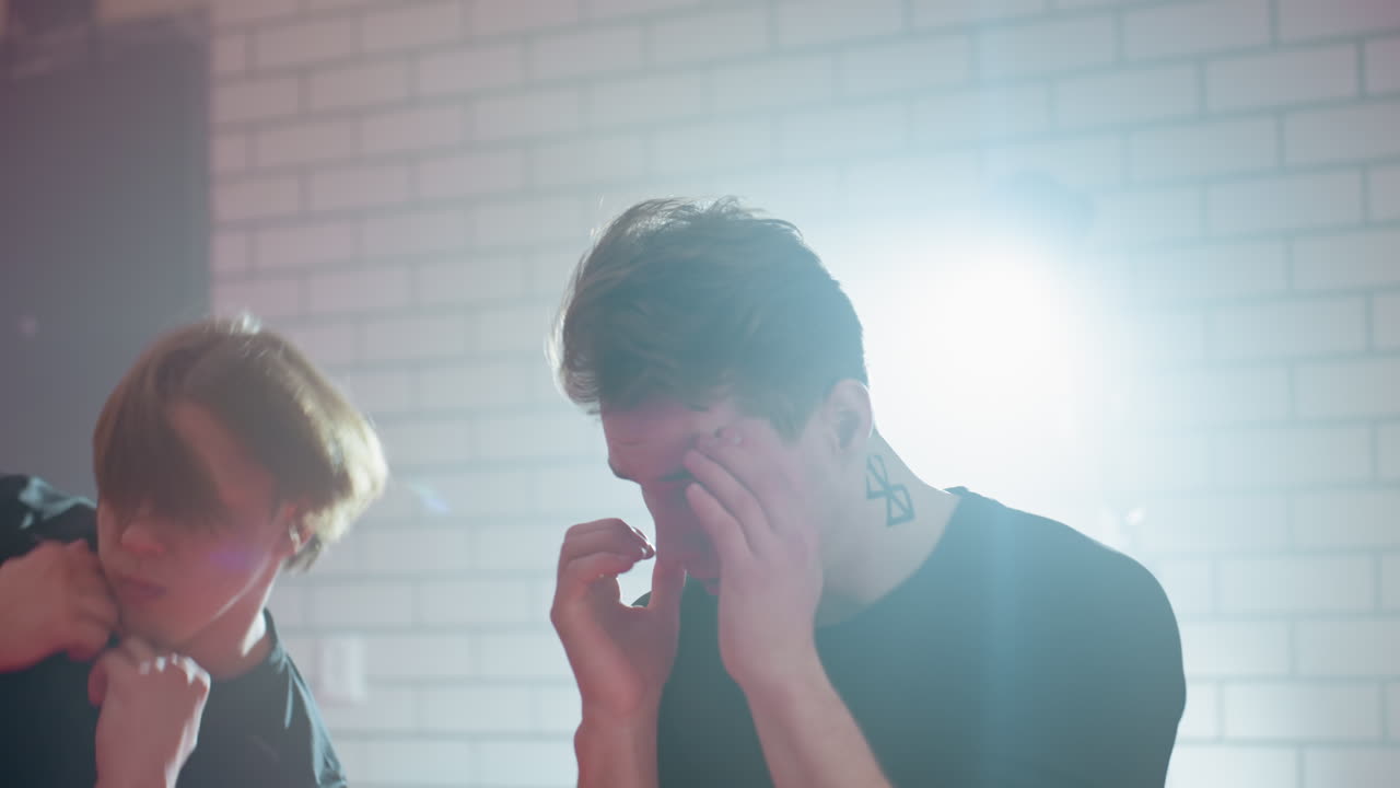 Two boxers in black shirts sparring indoors against white brick wall with bright backlight, practicing punches and defensive stances, showing intensity, focus, martial energy in training