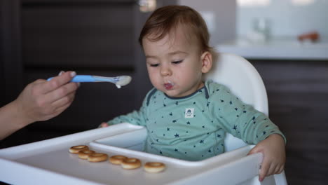 mommy is feeding the lovely baby sitting at the feeding table. mom gives full spoons to her little son. blurred backdrop.