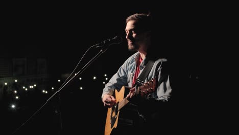 a musician is playing guitar on a large stage with audience members holding flashlights in the background. the auditorium is filled with beautiful flashing lights.