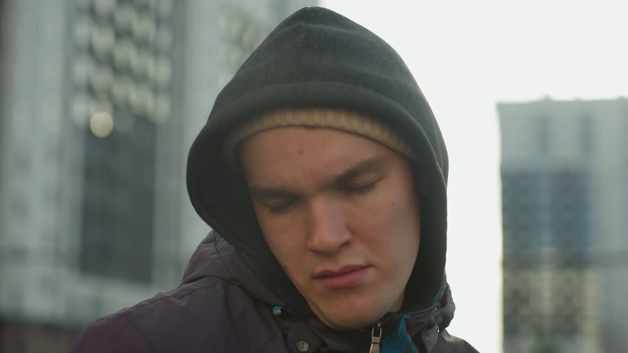 현대 주거 건물, 도시 환경 및 반사 포즈의 배경에 서서 생각 깊게 머리를 돌리는 후디와 베니를 입은 남자