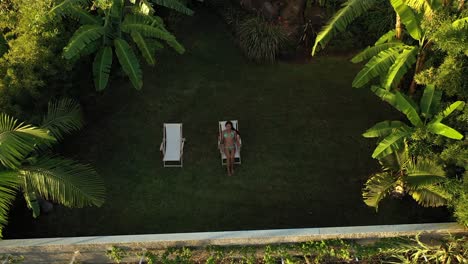 a drone shot of a young and beautiful woman sitting or laying down in the sunchair in a bikini