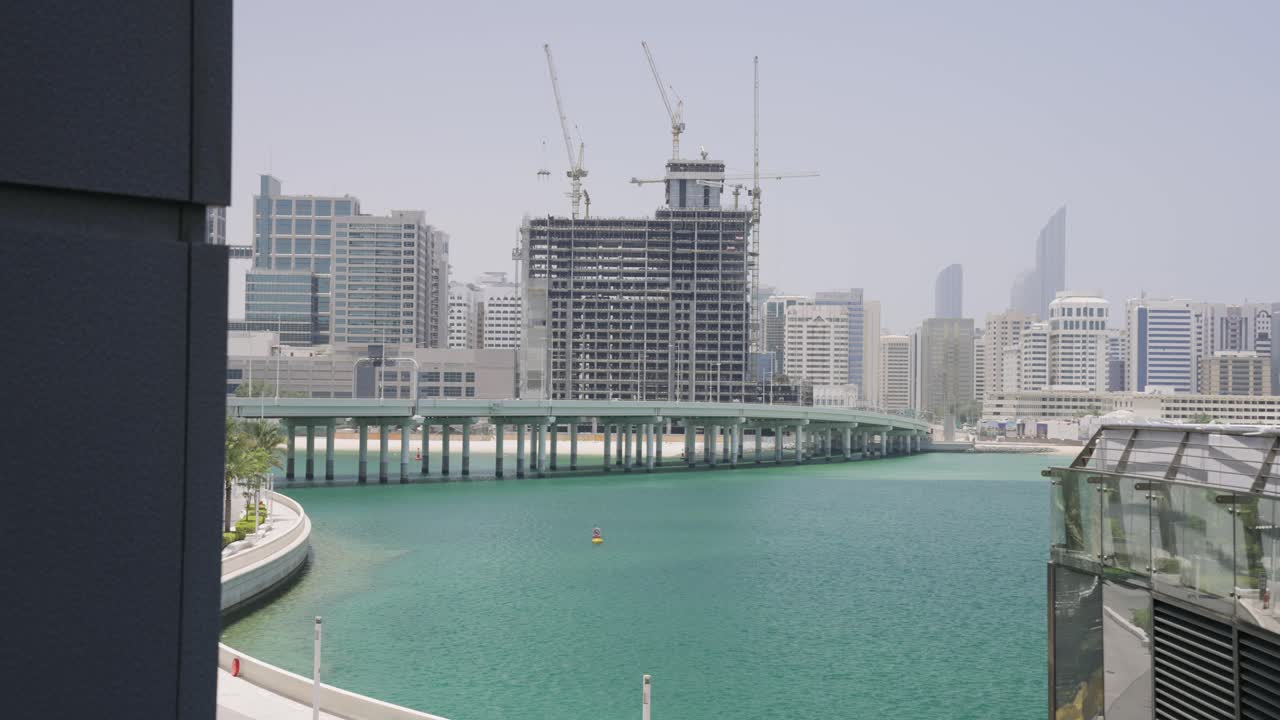Inquadratura cinematografica diurna ad angolo elevato che sovrasta il lago da un edificio ADGM, con costruzioni visibili che riflettono il rapido sviluppo di Abu Dhabi