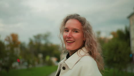 lady walking outdoors turns with gentle smile at camera curly hair fluttering in wind with blurred background showing soft evening light, street, greenery, and distant structures visible