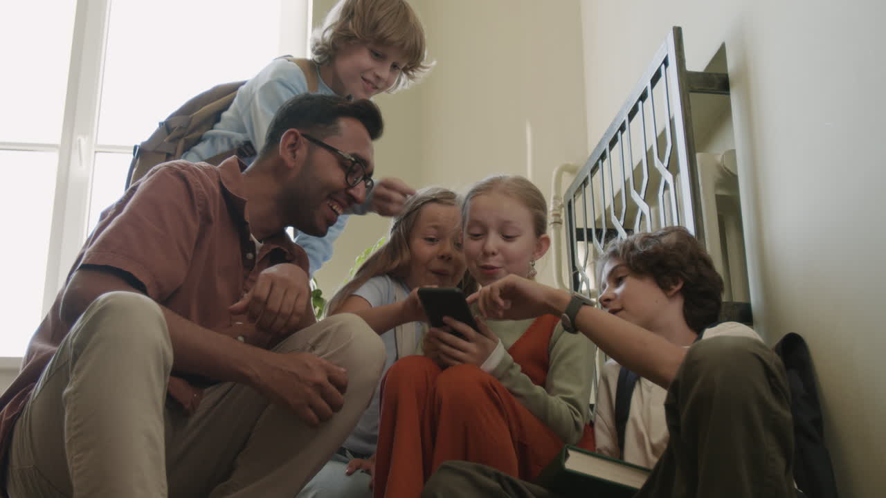 A diverse group of adults and children happily interacting and sharing moments, including using a smartphone, in a casual indoor setting