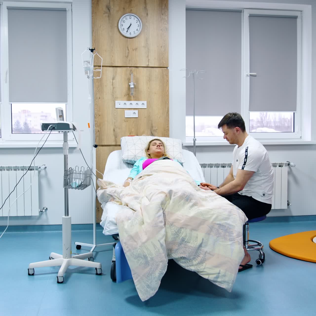 Loving husband sits at the bed of his pregnant wife. Blonde woman suffering from painful contractions. Modern hospital ward backdrop