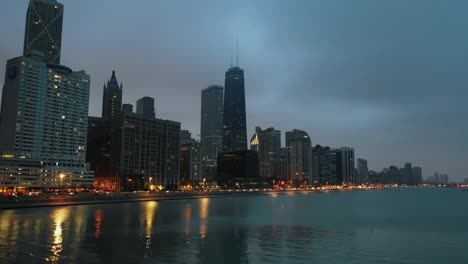 drone shot of city skyline with tall buildings and waterfront during evening in canada, with calm water reflecting lights from towers under cloudy sky, creating peaceful urban atmosphere