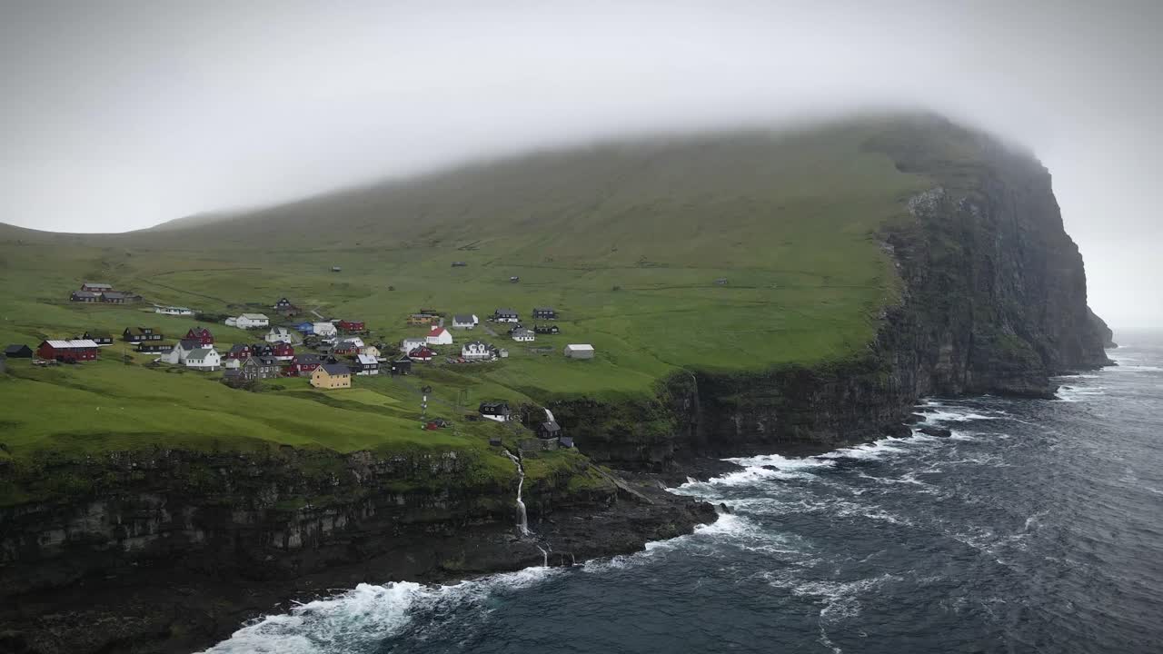 las nubes bajas que envuelven los dramáticos acantilados de la isla de kalsoy, parte de las islas feroe, revelan olas que se estrellan contra la costa rocosa debajo de un pintoresco pueblo en la ladera de una colina.