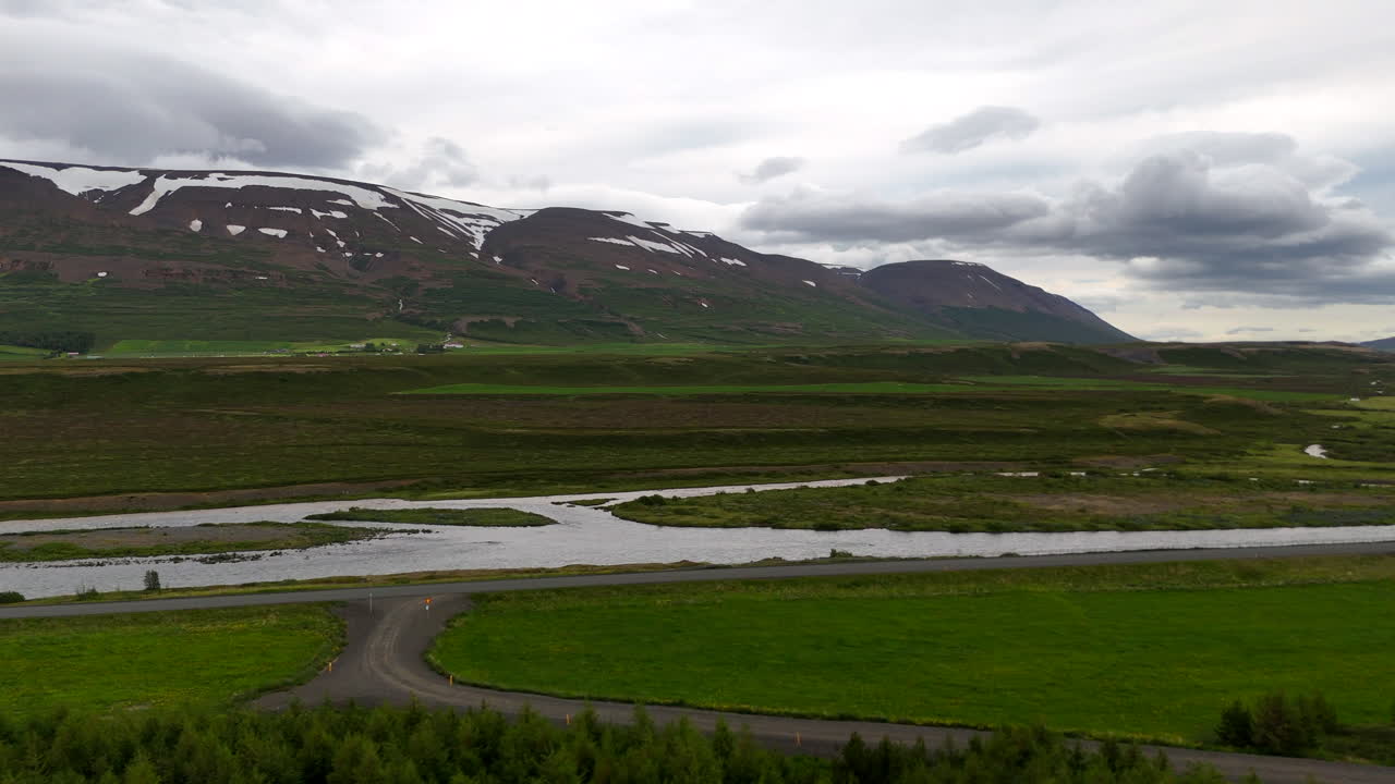 Aerial view of the Skjálfandafljót River winding toward Skjálfandi Bay, framed by broad green valleys, scattered farms and snow-marked mountains under a dramatic sky in northern Iceland