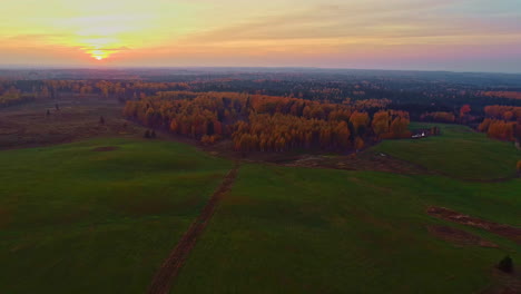 retroceso aéreo de la puesta del sol sobre vastos prados verdes junto al paisaje boscoso con dosel en colores otoñales - hermosa escena de la hora dorada con horizonte naranja
