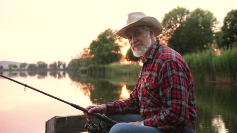 portrait of a senior fisherman wearing hat and sitting on the lake pier while holding a fishing rod at sunset