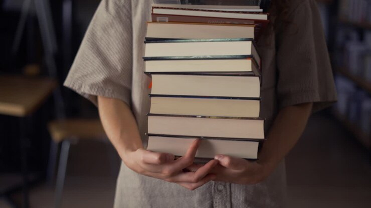 Estudiante irreconocible camina con muchos libros en la biblioteca, preparándose para los exámenes
