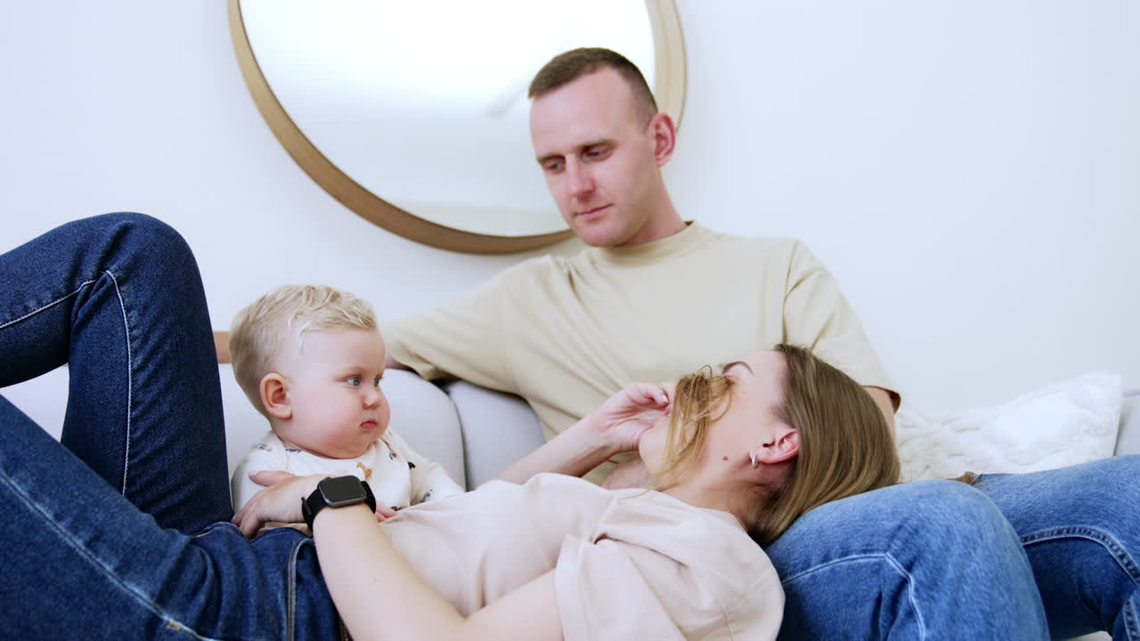 Mommy hides her face behind her hair from a baby. Smiling kid uncovers mom's face. Father is watching smiling. Family time together.