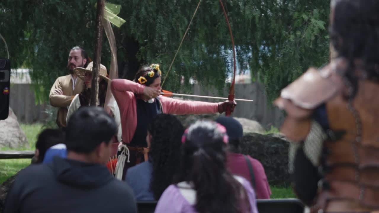 A group of people attend an outdoor medieval cultural event with costumed participants, a rustic setting surrounded by greenery.