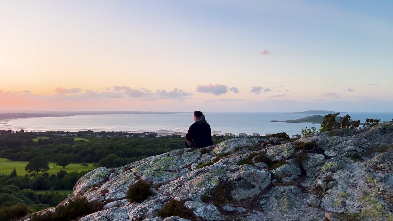 Handheld shot of man sitting on rocky mountaintop watching sunset with Ireland’s Eye island in the distance