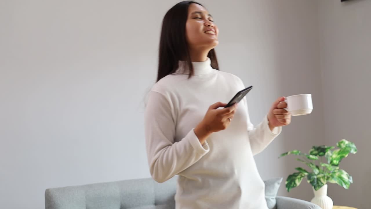 Donna asiatica felice e spensierata che tiene in mano una tazza di caffè e uno smartphone, si diverte muovendosi ascoltando musica e si rilassa in soggiorno.