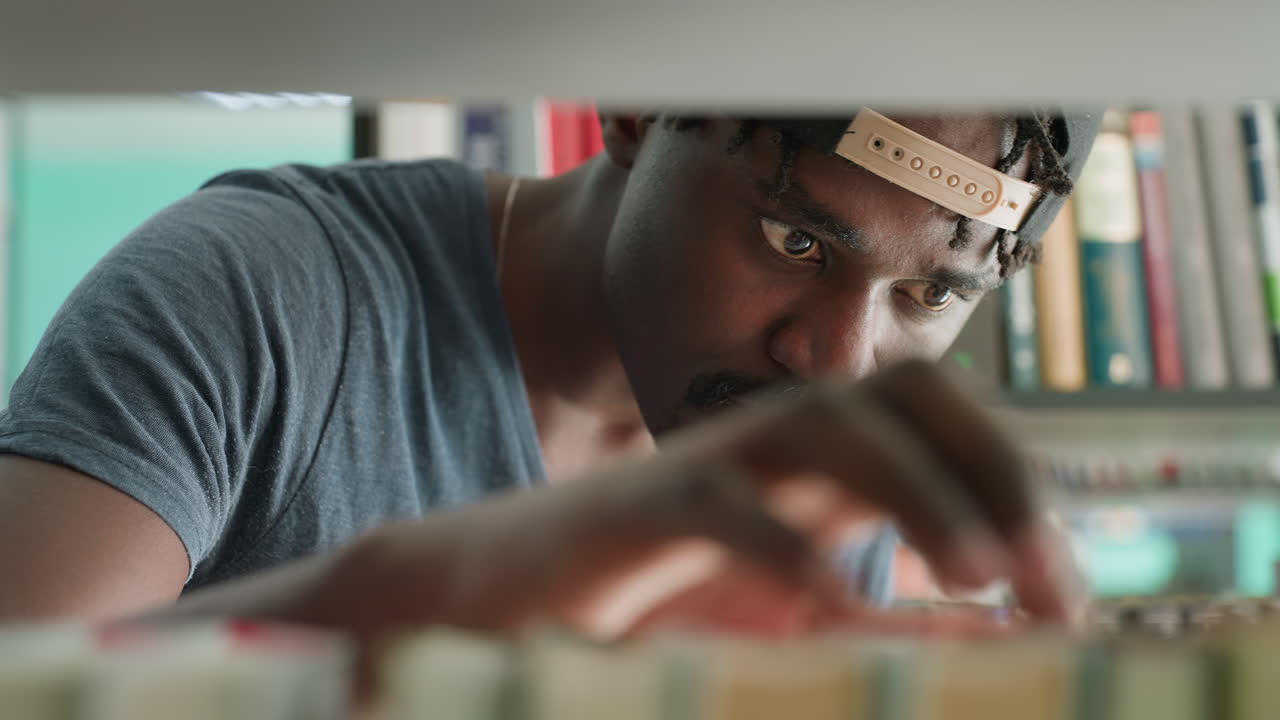 close up from shelf shows student in cap gliding hand across arranged books, eyes intent through gap, browsing titles in quiet library aisle with teal walls, soft daylight