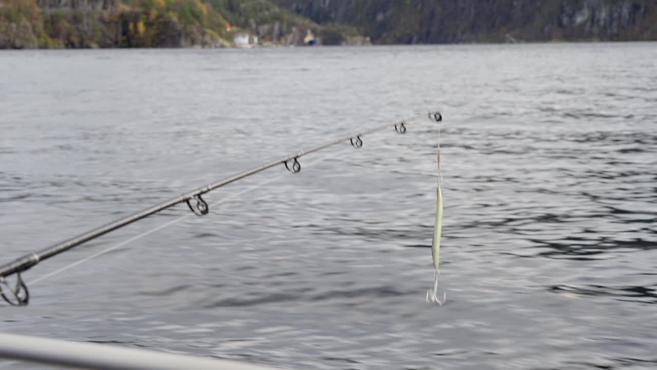 Heavy fishing lure and solid rod hanging over the water from a moving boat, with sea surface below