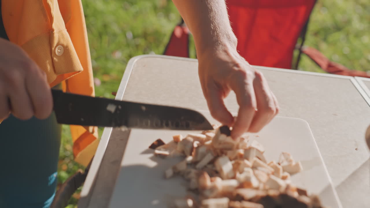 Manos preparando la mesa de camping, rebanando champiñones sobre una tabla de cortar de plástico, con un fondo de césped brillante y una silla roja. Un ambiente diurno y casual, con trozos bien picados recogidos en un tazón, mostrando una técnica de cuchillo firme y amigable.