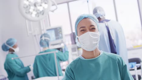 Video-portrait-of-biracial-female-surgeon-in-mask-smiling-in-operating-theatre,-copy-space