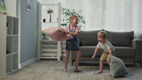 girl hits younger brother with pillow standing near sofa