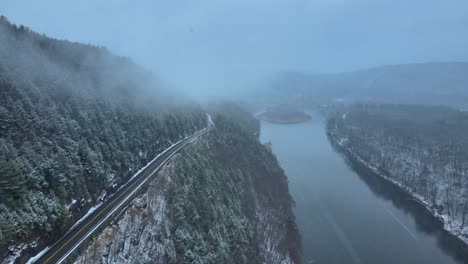 Aerial-footage-of-a-snowy,-scenic-byway,-winding-mountain-valley-road-during-a-snowstorm-with-pine-trees,-a-river,-mountain-highway,-rocky-cliffs,-and-forests-during-winter-on-a-cold,-blue-day