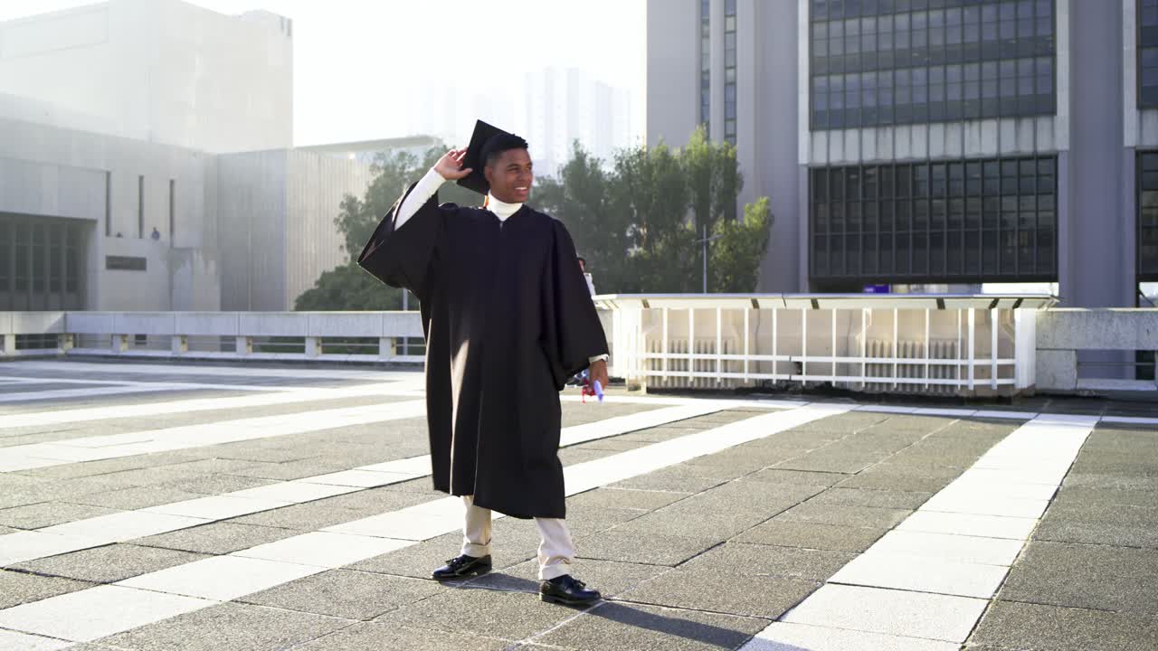 Male graduate wearing gown and cap holding diploma, swinging it, jumping in celebration on plaza